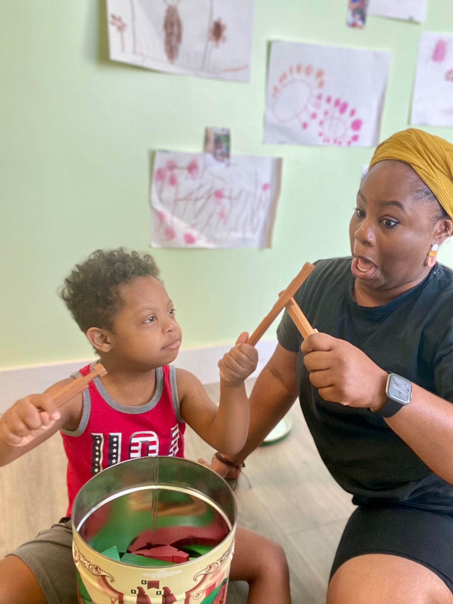 Bintu and a child holding wooden rhythm sticks beside a drum