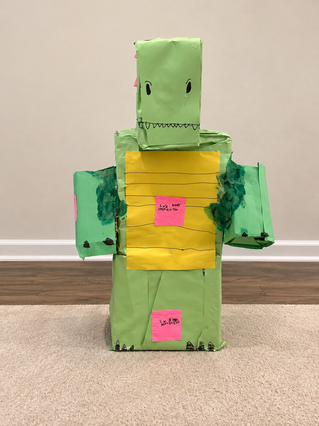 Completed green cardboard dinosaur with pink handwritten volume-calculation labels against a cleaned background